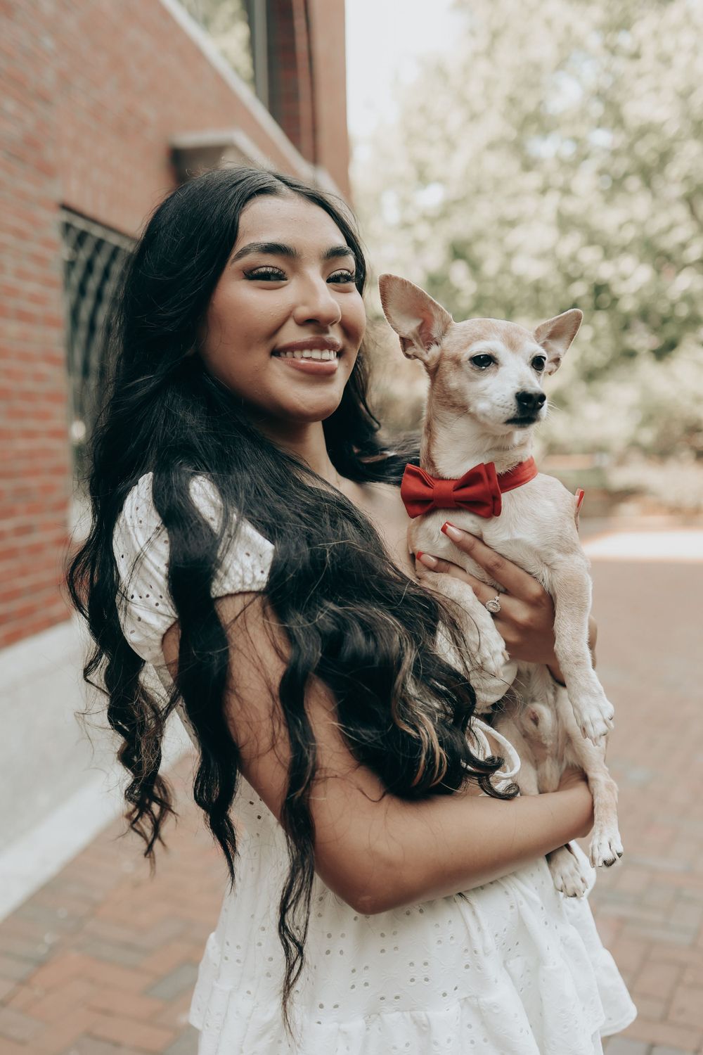 Graduate in white dress holds small dog wearing red bowtie against brick building backdrop.