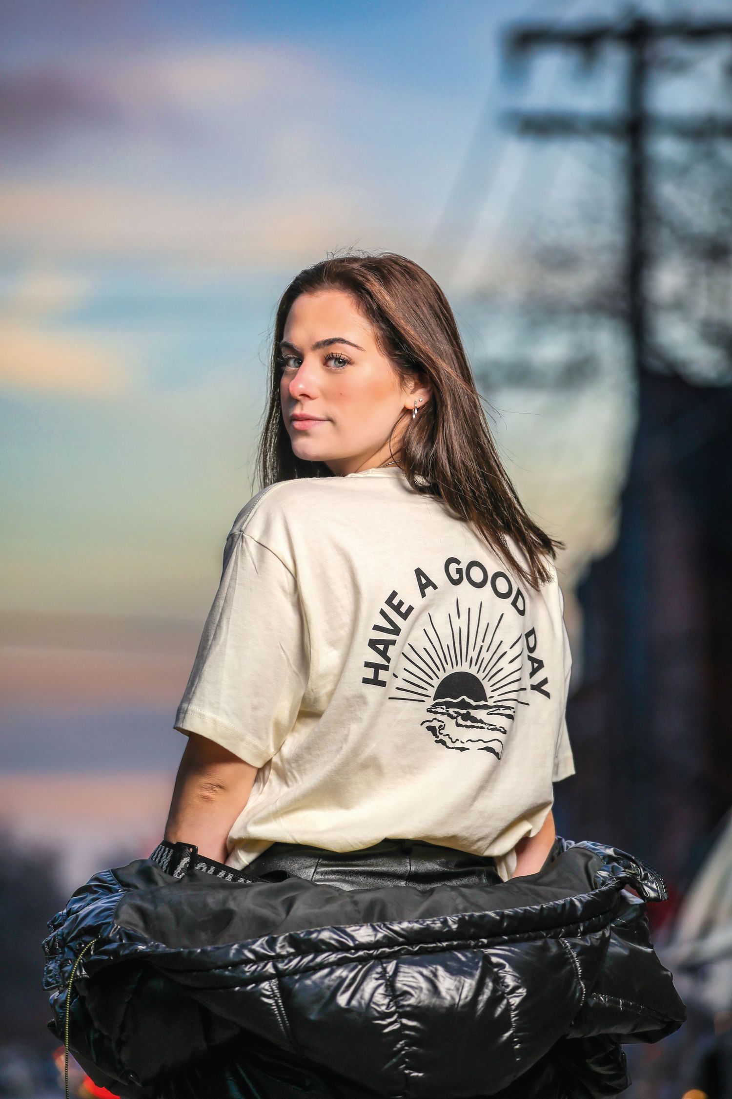 Young woman in a cream Have A Good Day t-shirt holding a black jacket at an outdoor festival at sunset.