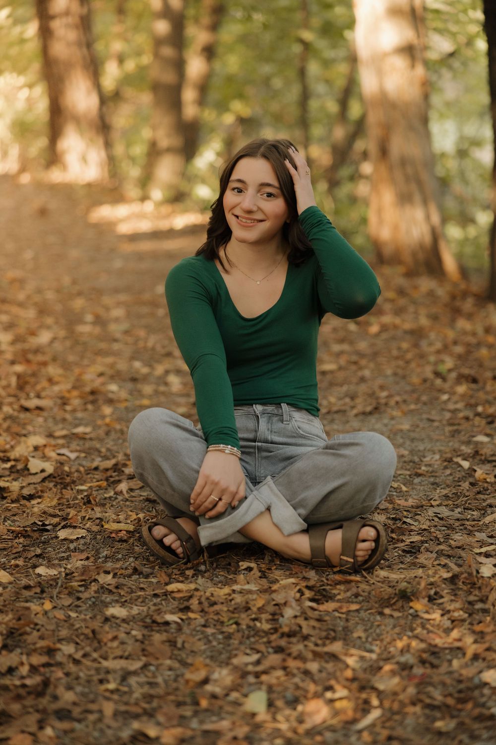 A person in a green sweater sits cross-legged on autumn leaves in a wooded setting.