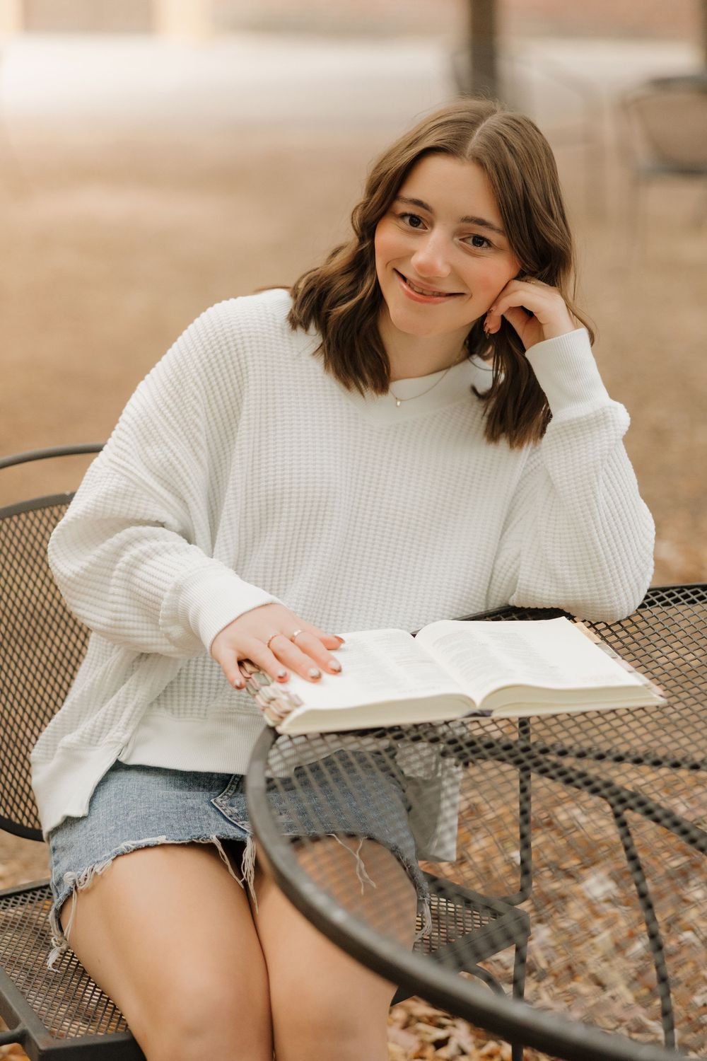 Student in cozy white sweater reading a book while sitting at outdoor metal patio table.