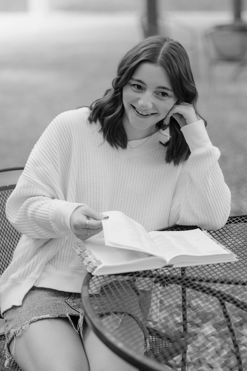 A person in a white knit sweater sits at a patio table reading a book with a warm smile in black and white.