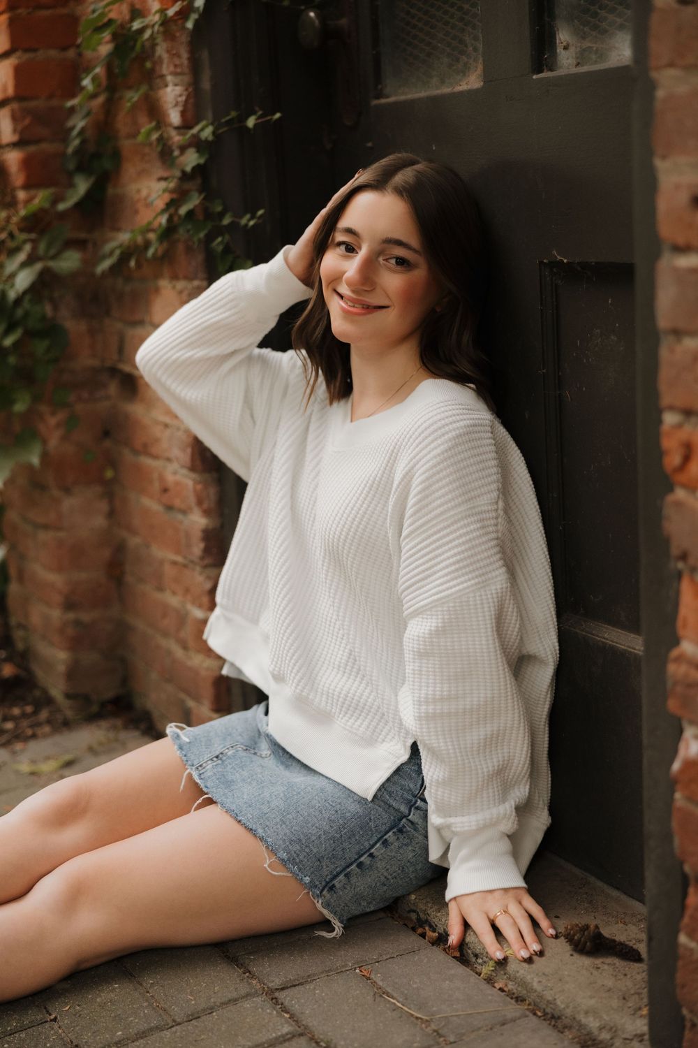 Person in white knit sweater and denim shorts posing casually against brick wall background.