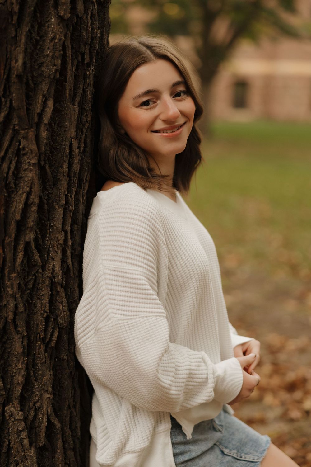 Autumn outdoor photoshoot with white knit sweater against tree backdrop with fallen leaves.