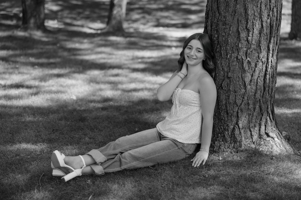 Woman in white dress sitting against tree trunk in outdoor setting with dappled sunlight.