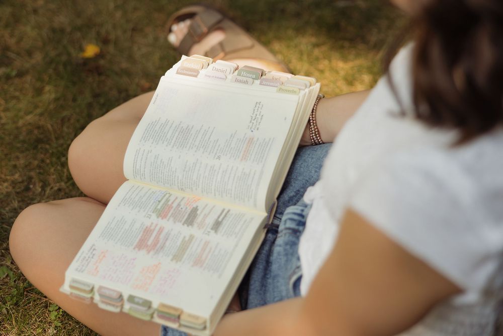 A person sitting in grass reading a book with pages open on their lap in natural outdoor lighting.