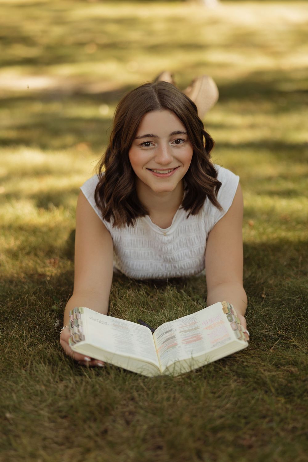 Someone lies on grass while reading a book outdoors on a sunny day, wearing a white sleeveless top.