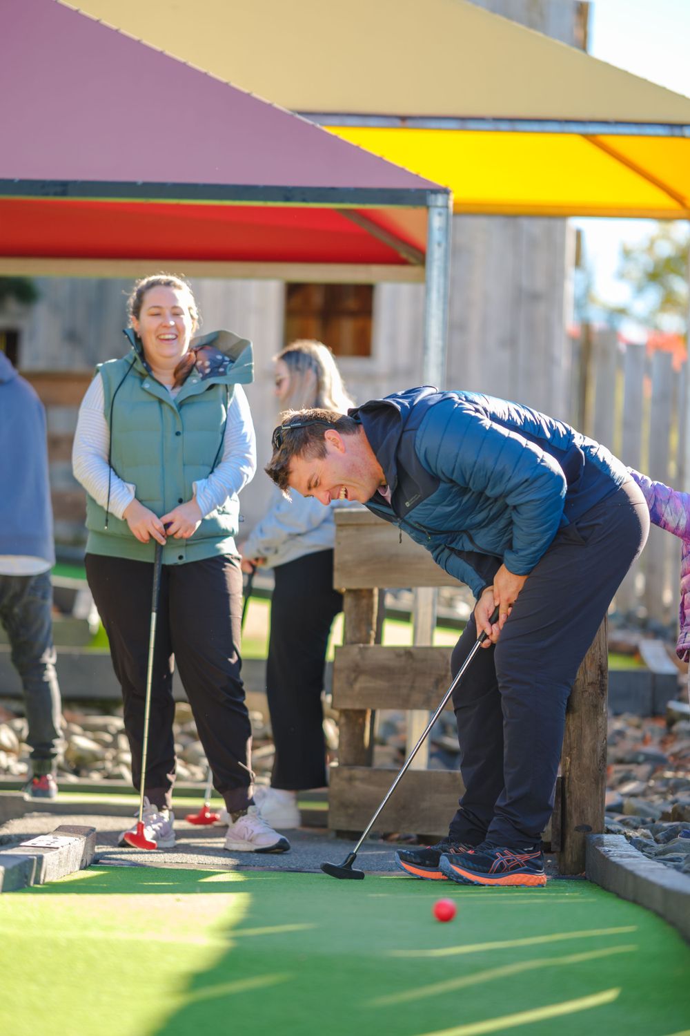 People bent over putting on a miniature golf course with bright colored awnings overhead.