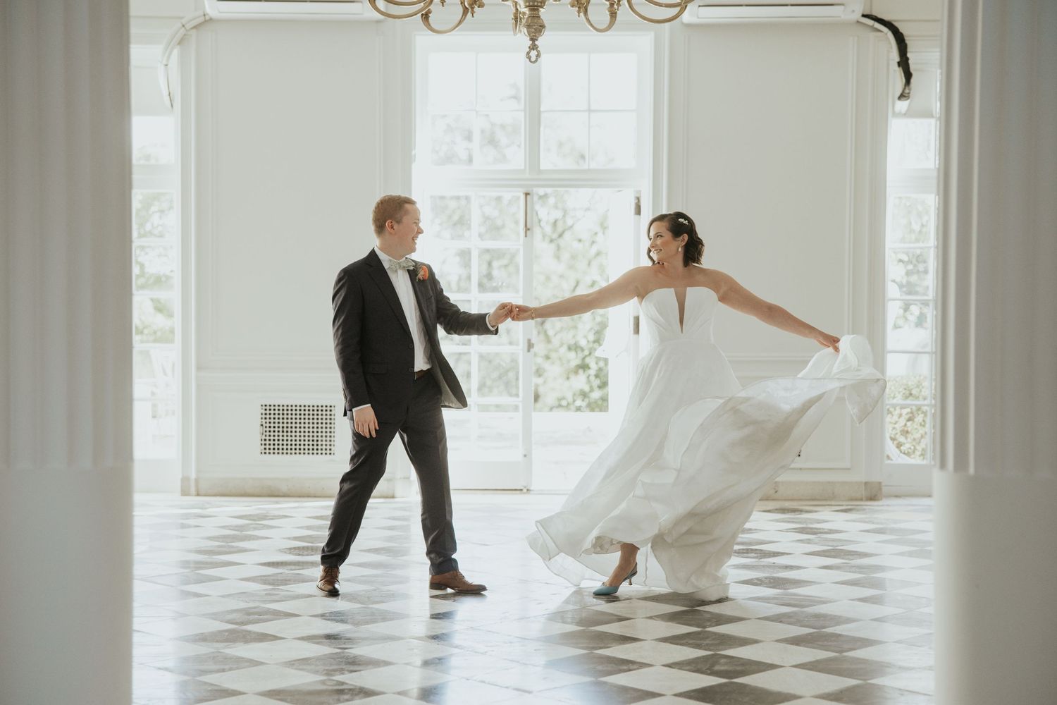 A couple dancing together in an elegant white room with checkered floors and large windows.