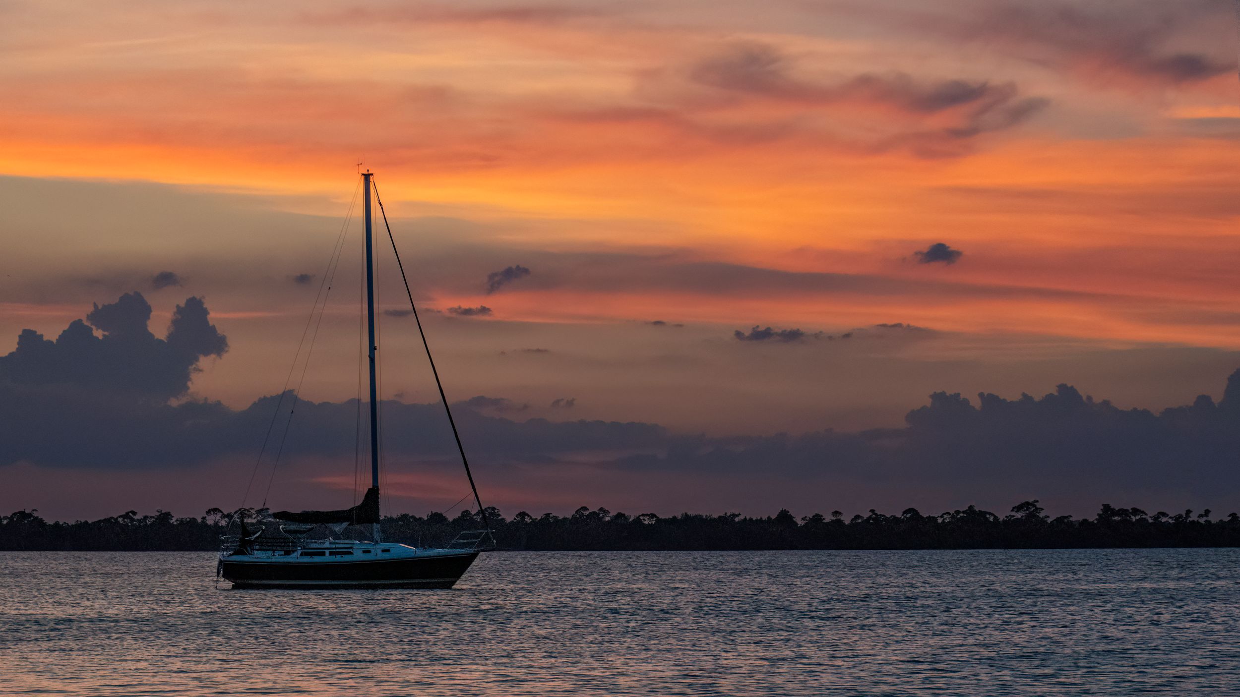 Silhouette of a sailboat against a dramatic orange and purple sunset over water.