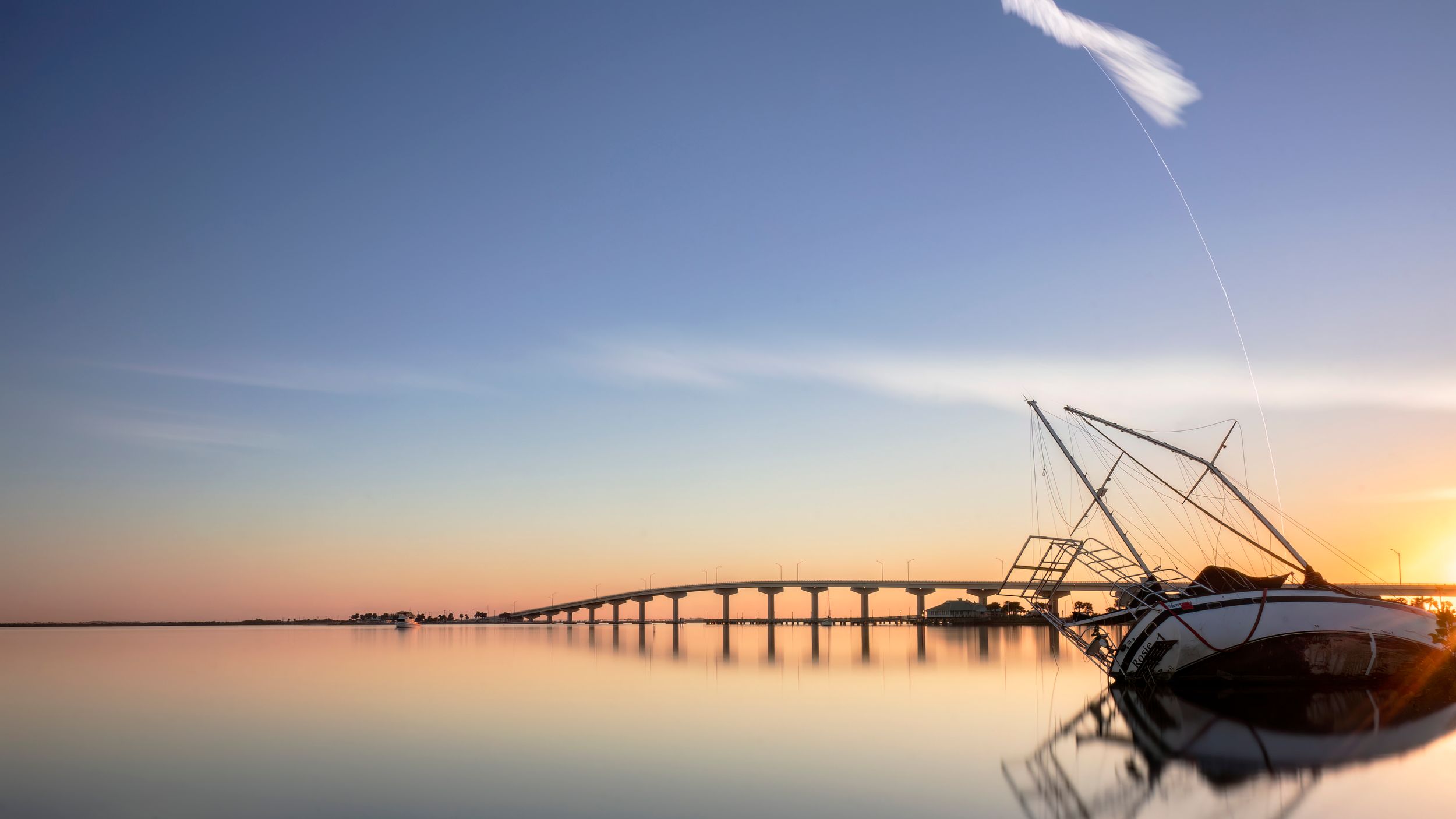 Panoramic coastal landscape with bridge silhouette and fishing boat at sunset.