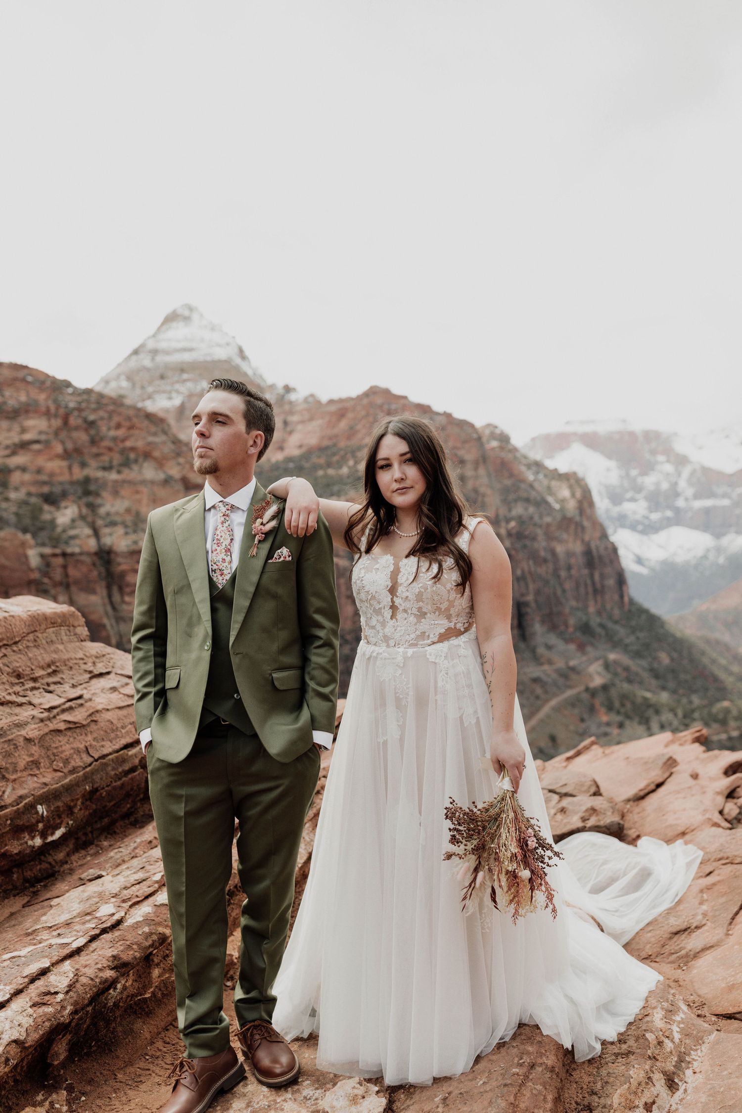 Wedding couple poses on red rock cliffs with snow-capped mountains in the background in Zion National Park.