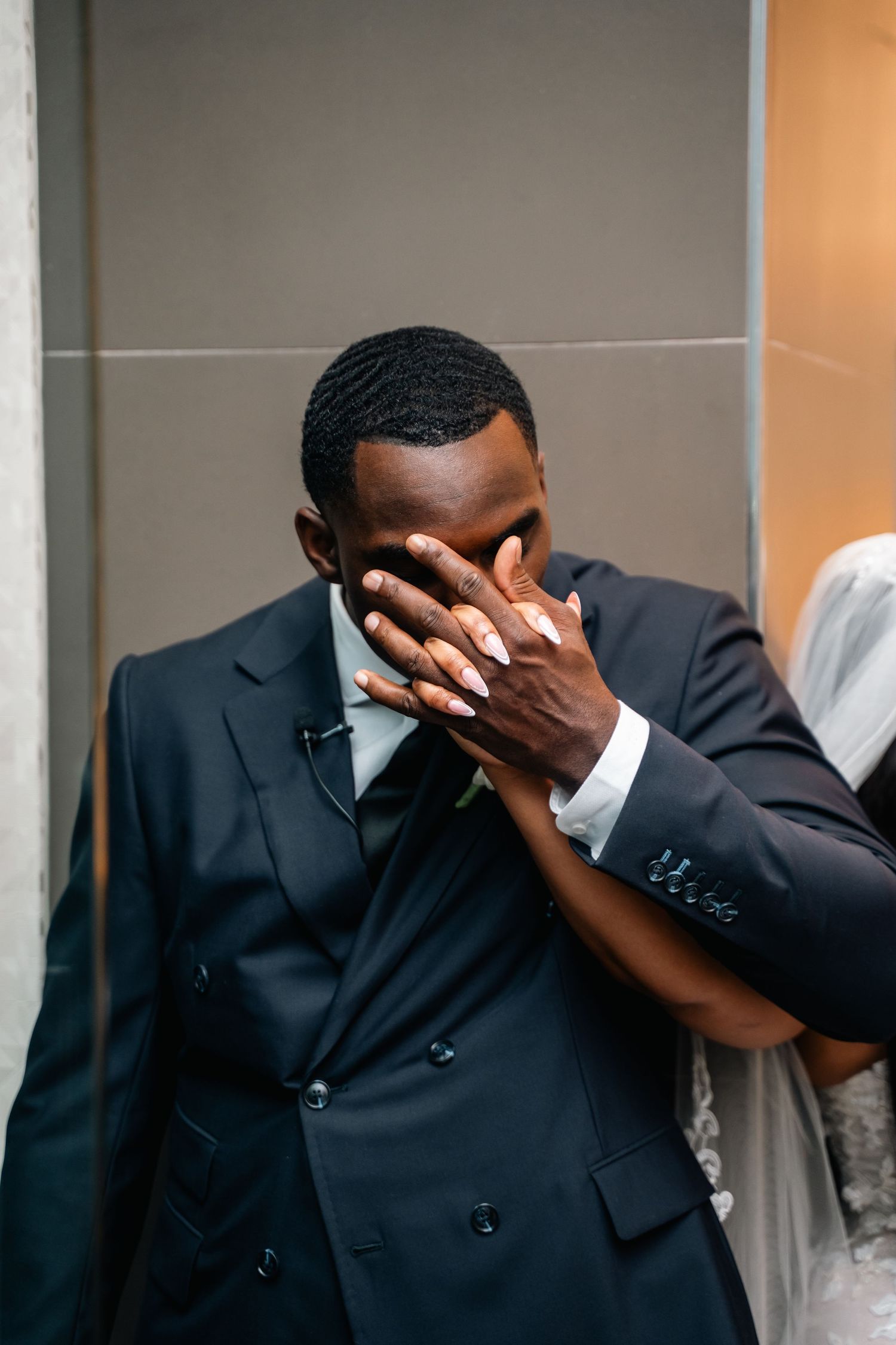 Emotional groom in black suit covers face with hands during wedding moment.