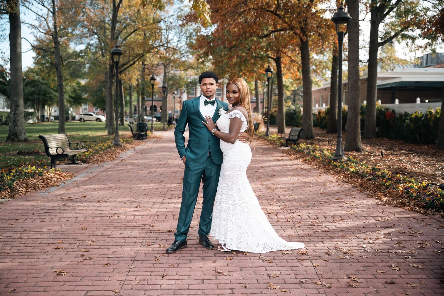Elegant couple in wedding attire pose on brick pathway during fall season.