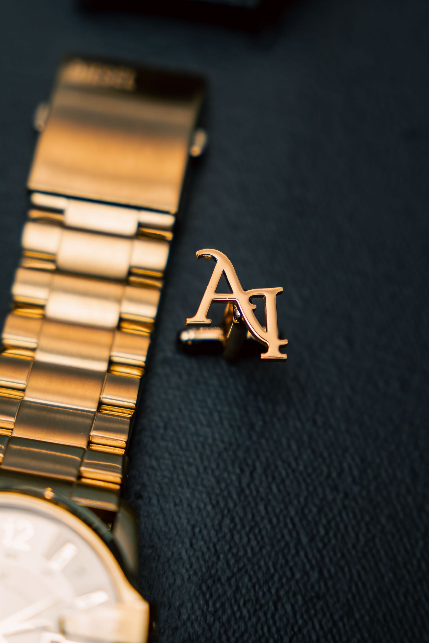 Gold watch band and matching 'AI' monogram pin on dark textile background.