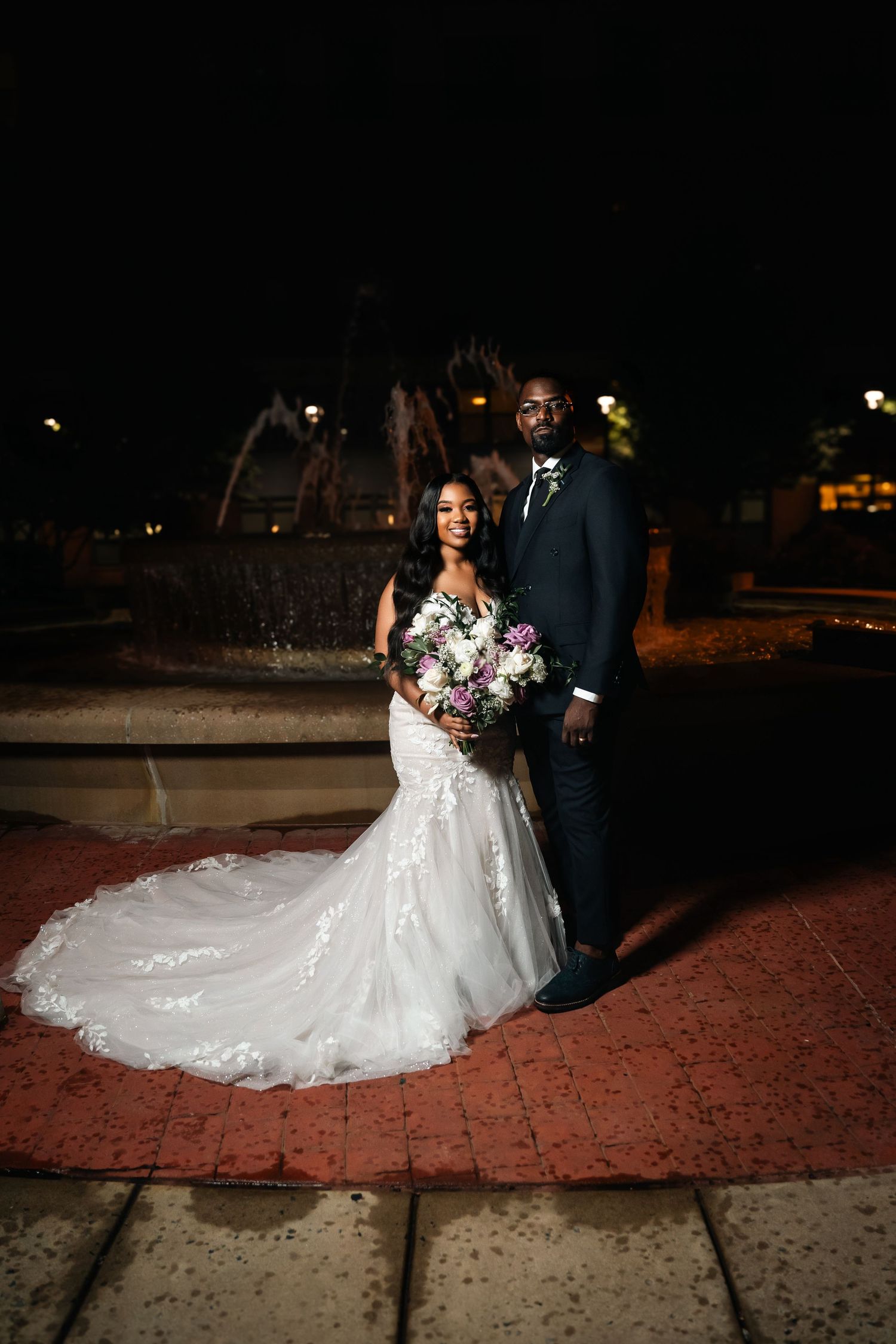 Romantic nighttime wedding portrait by fountain with bride holding floral bouquet.