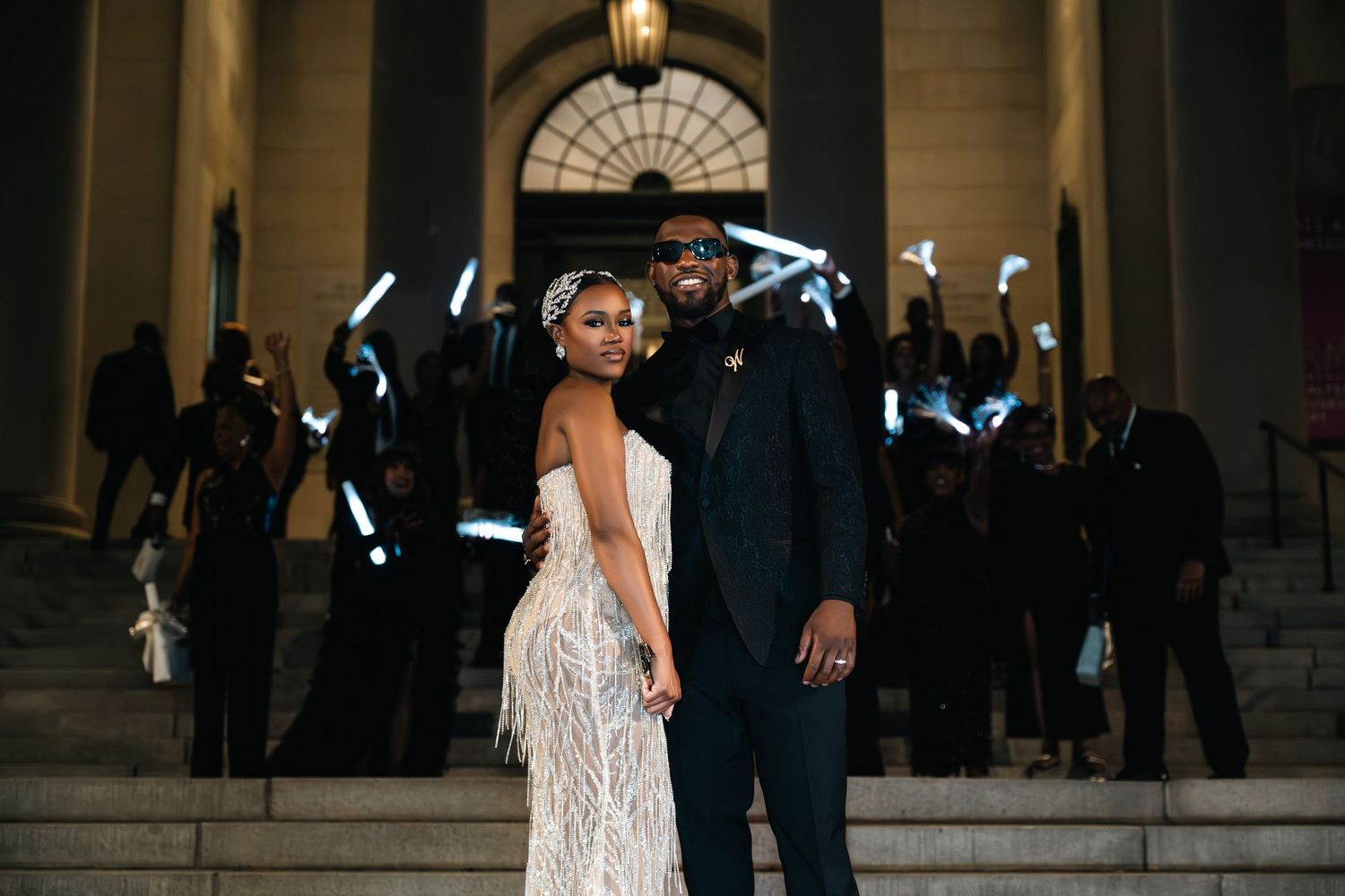 Glamorous couple poses on steps at night with illuminated backdrop.