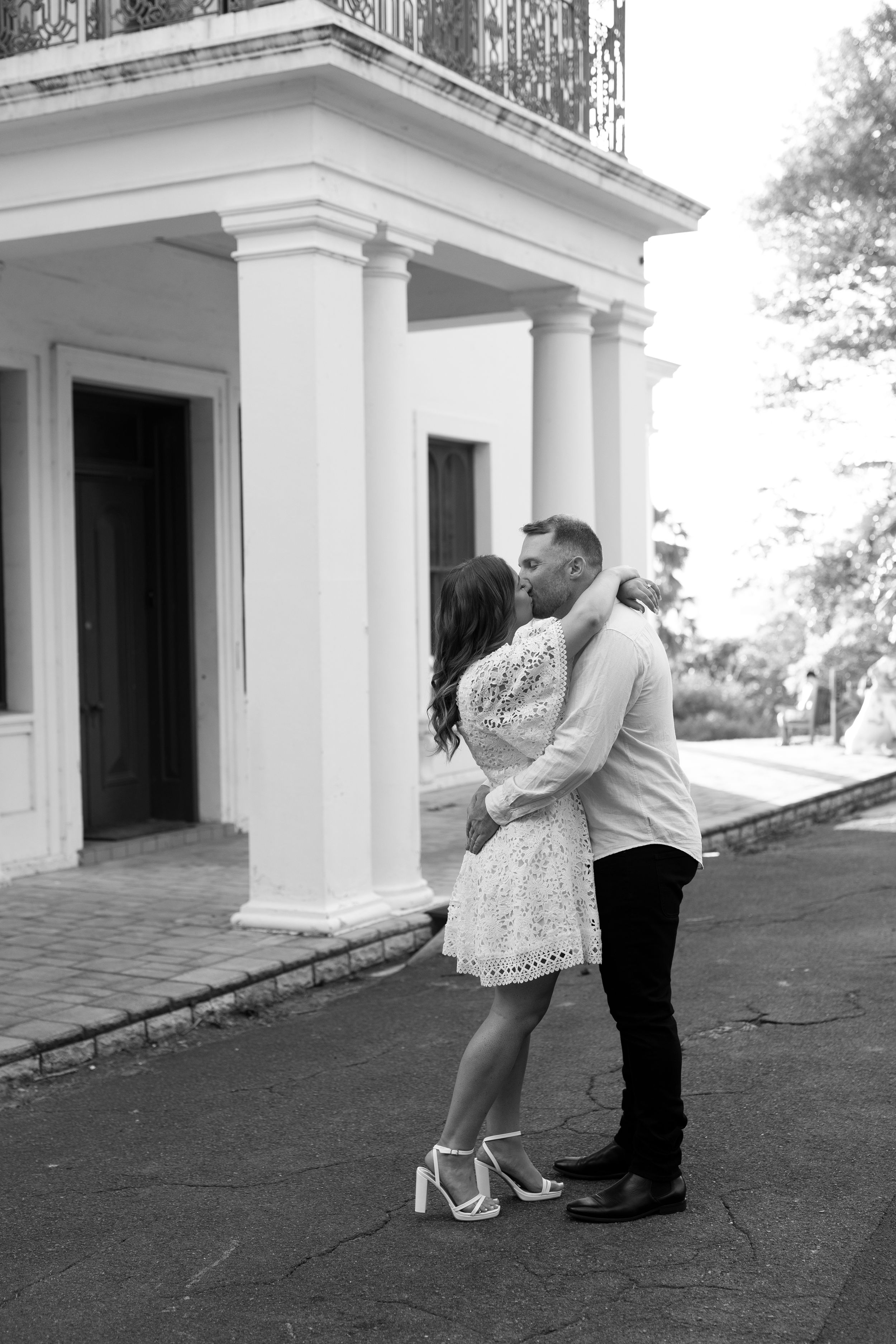 A romantic couple shares an embrace on the porch of a white columned building in black and white photography.