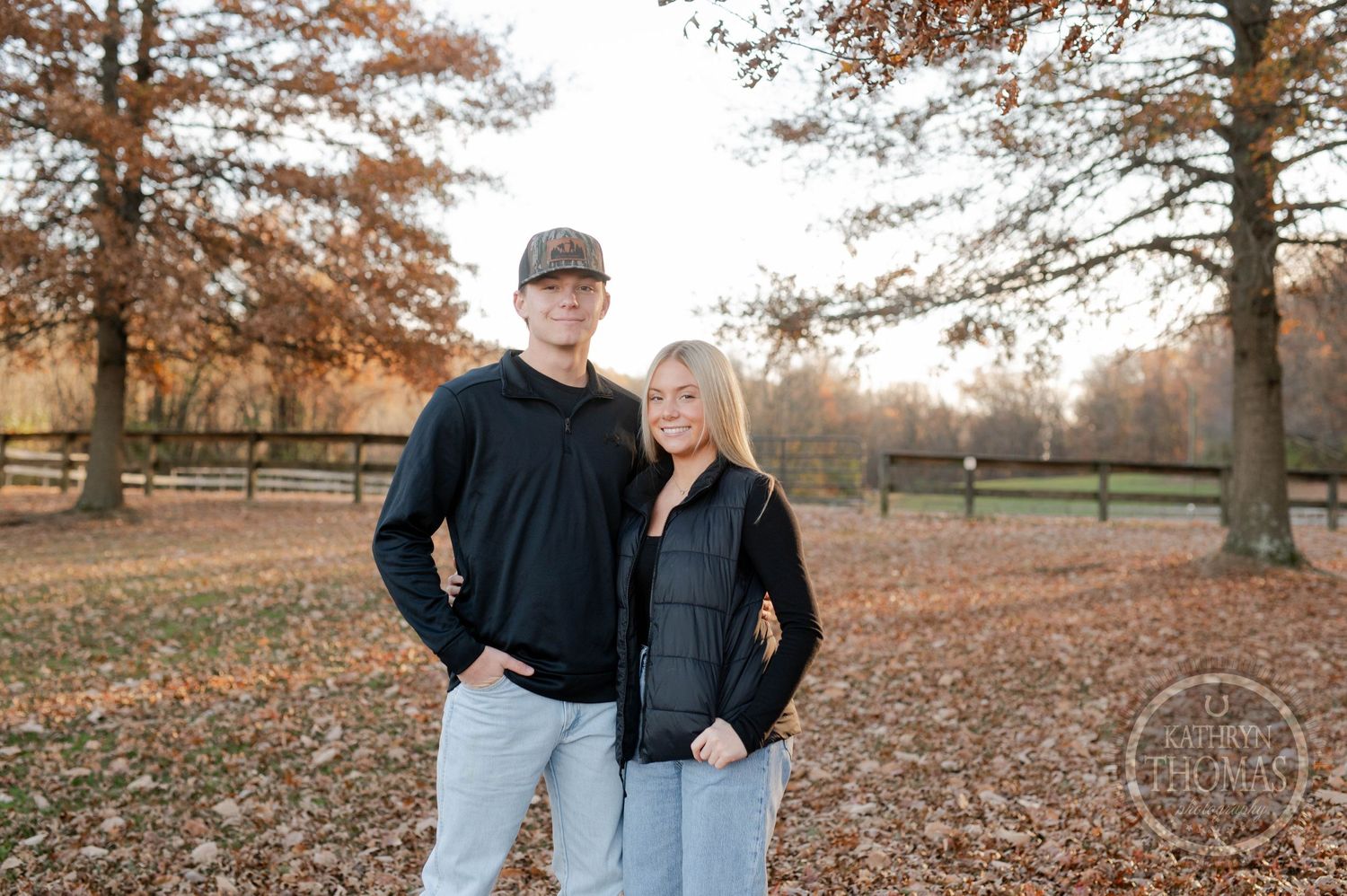 older siblings  in coordinating black outfits poses in front of autumn trees and wooden fence.