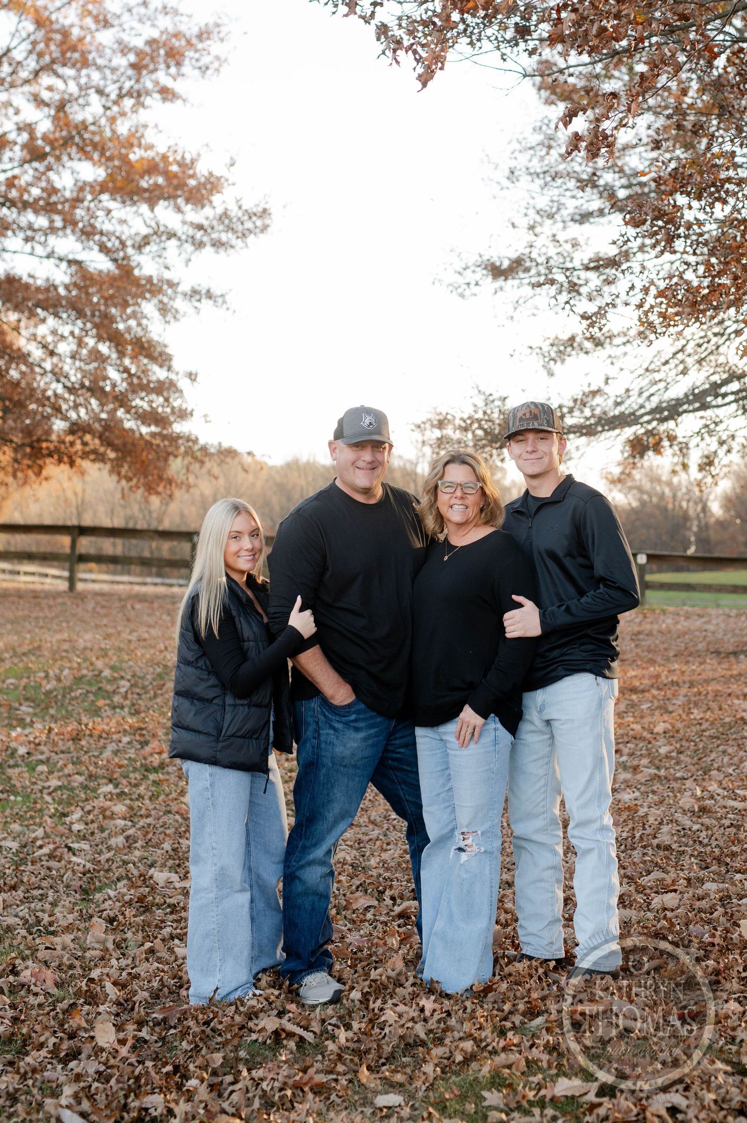 Group in casual attire poses together on fallen leaves near wooden fence during autumn.