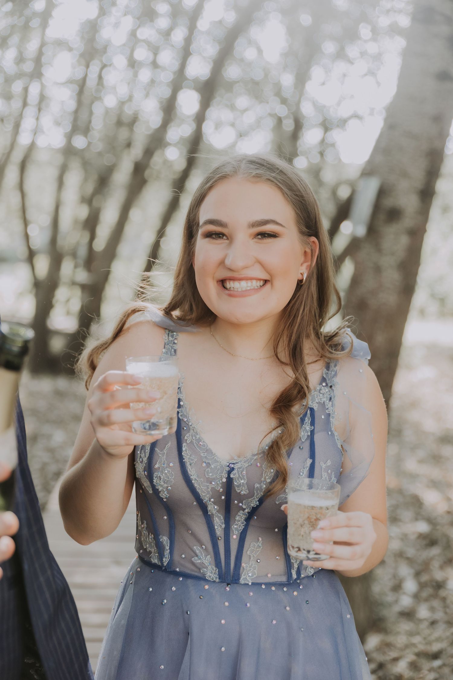 A person in a sparkly blue prom dress holding drinks and smiling in a forest setting.