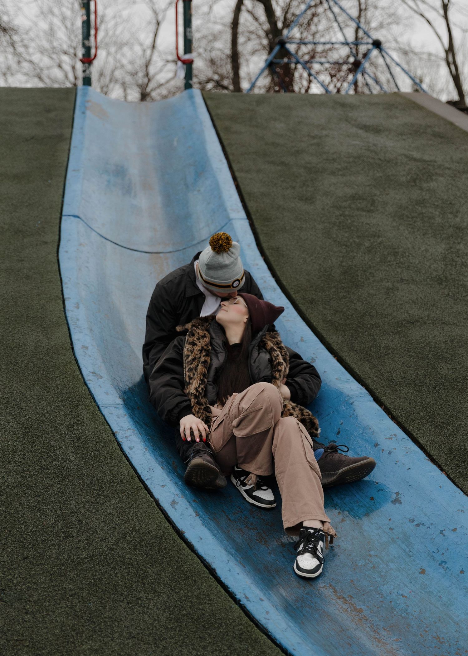 A person in winter clothing lies on a blue playground slide on a chilly day.