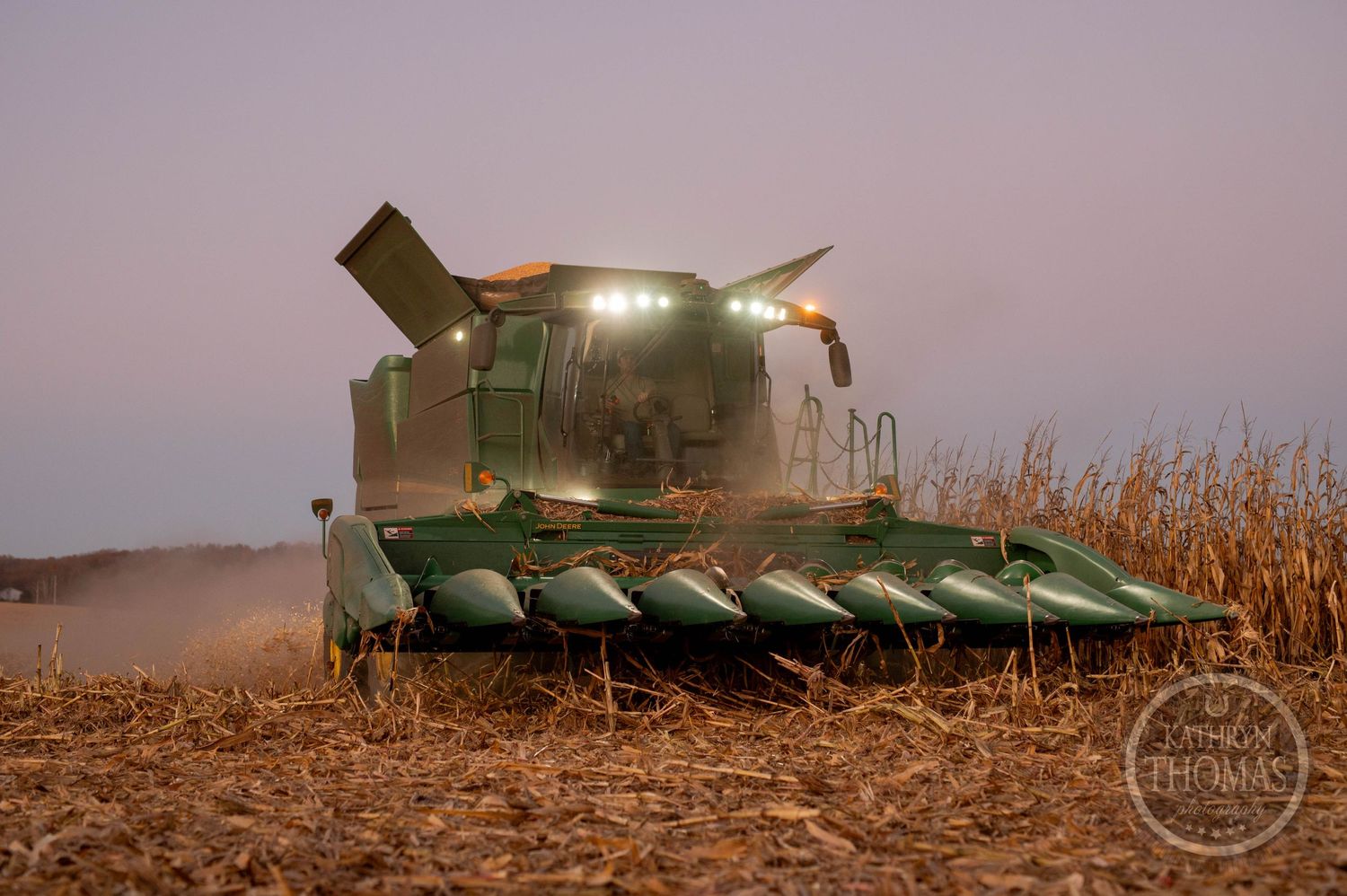 Green combine harvester with corn head attachment working through a cornfield at dusk.
