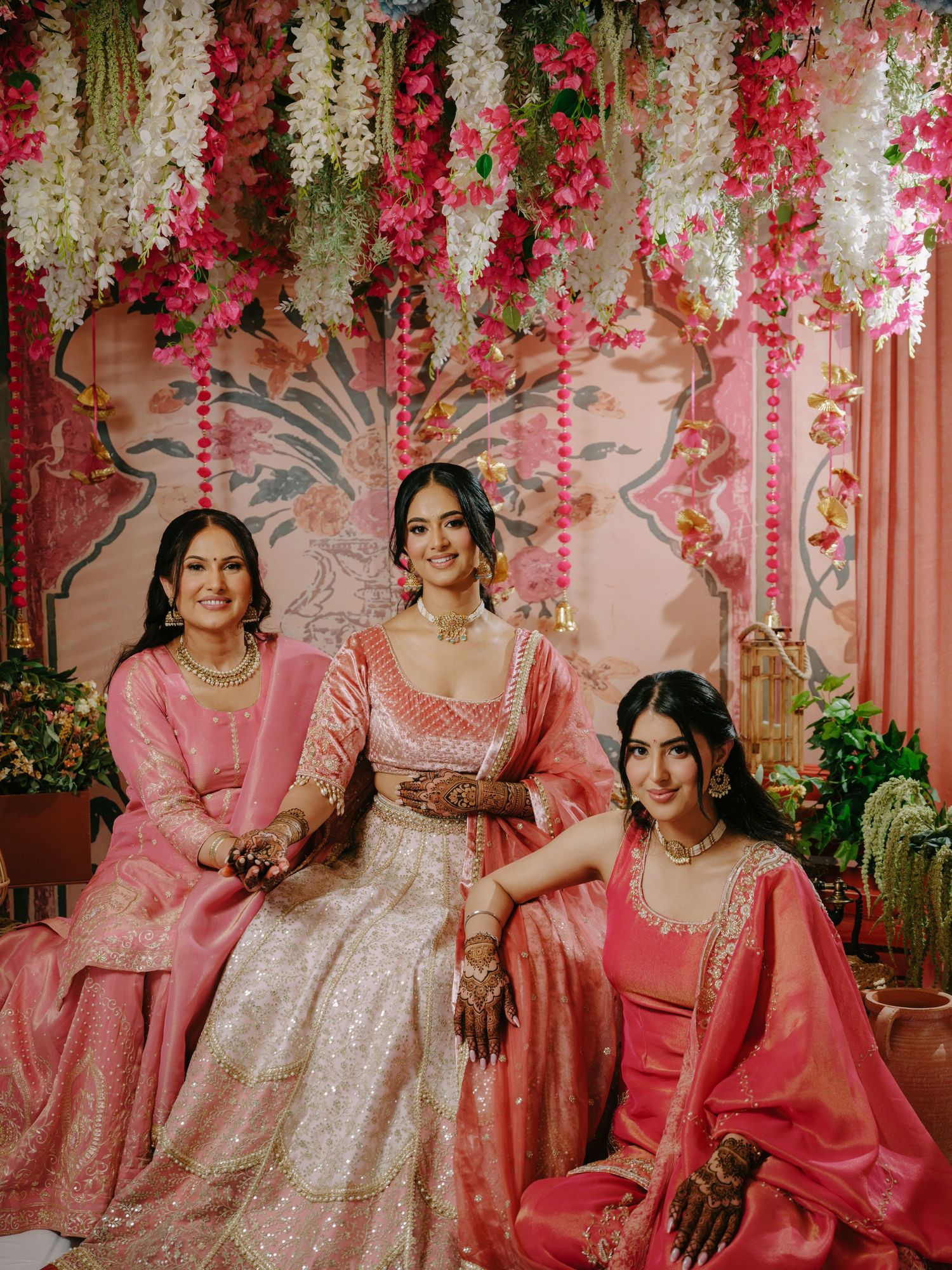 Decorative floral backdrop with hanging pink and white flowers frames three people in elegant traditional pink attire.