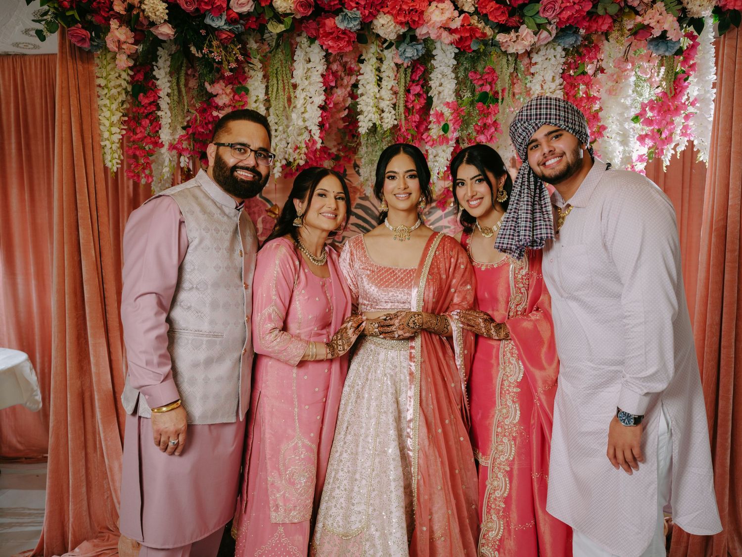 A group in coordinated pink and cream traditional Indian attire poses together at a decorated floral wedding backdrop.