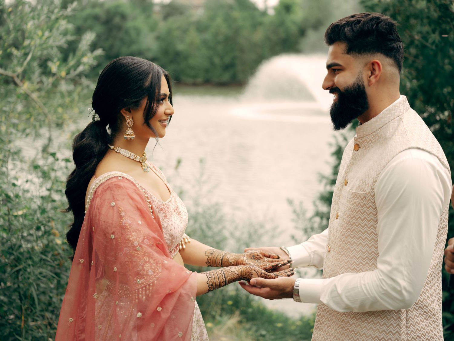 A couple in traditional South Asian attire holds hands outdoors surrounded by greenery during a romantic moment.
