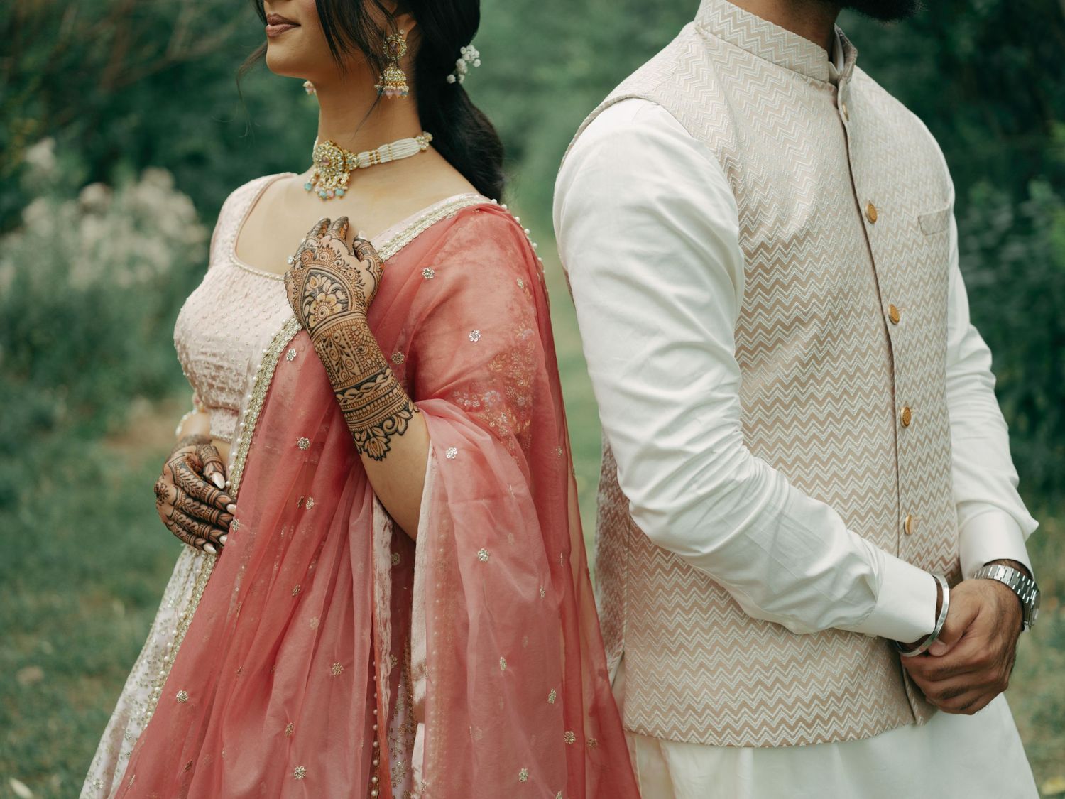 A couple in traditional wedding attire with coral pink sari and white embroidered sherwani pose outdoors together.