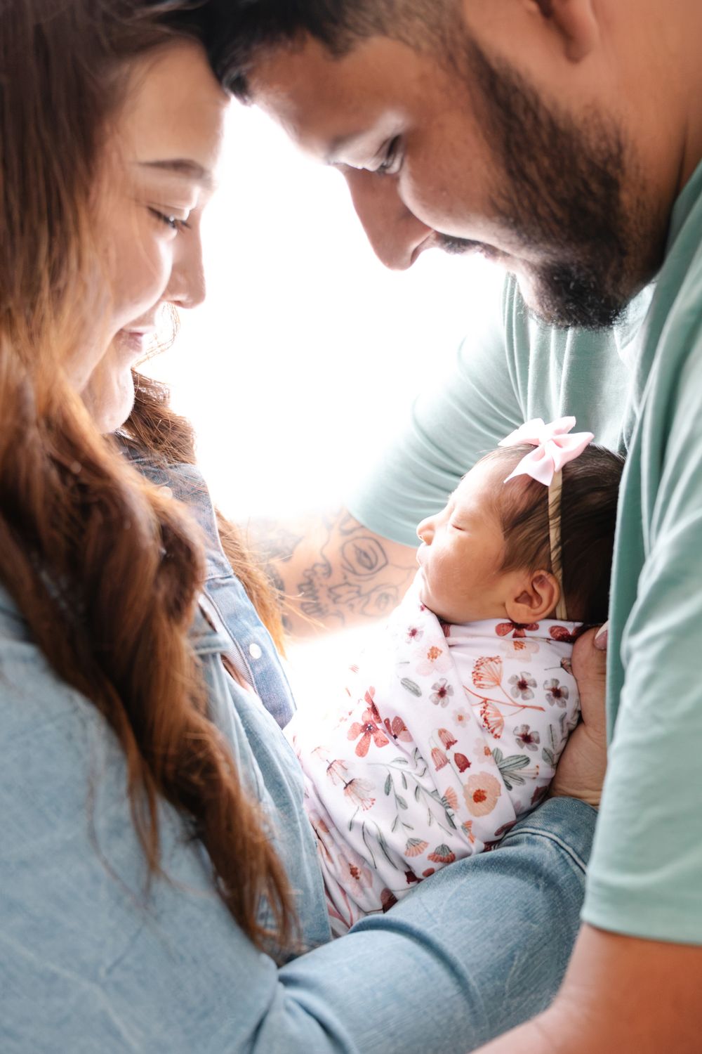 New parents lovingly gaze at their swaddled newborn baby girl wearing a pink bow headband.