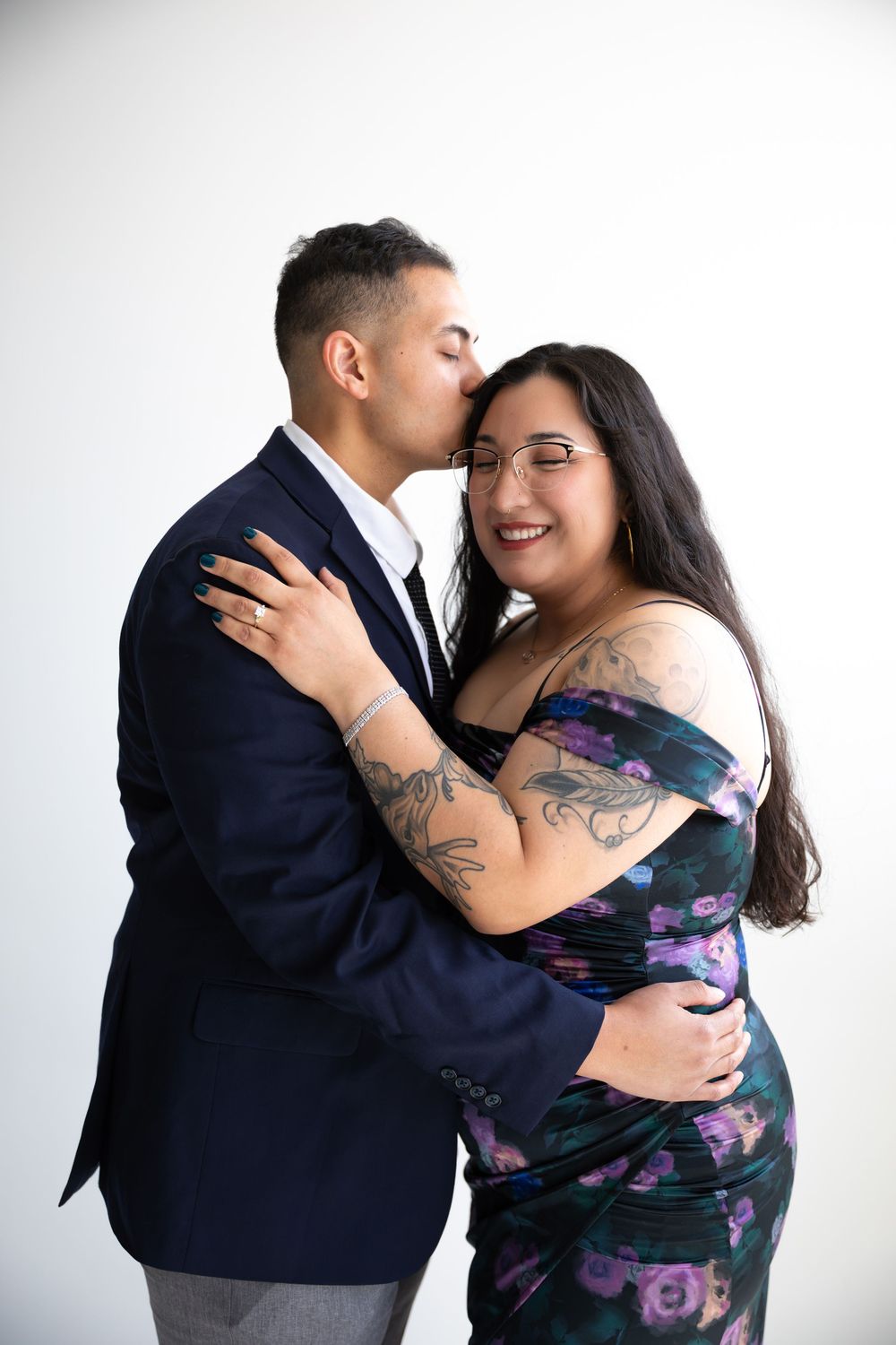 A couple embraces and shares a kiss, with the woman in a floral off-shoulder dress and the man in a navy suit.