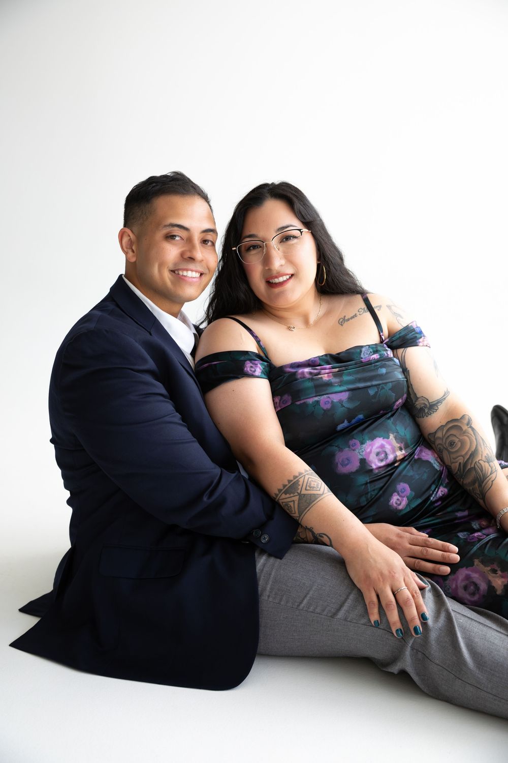 A smiling couple poses together on a white studio floor, man in navy blazer, woman in floral dress with tattoos.