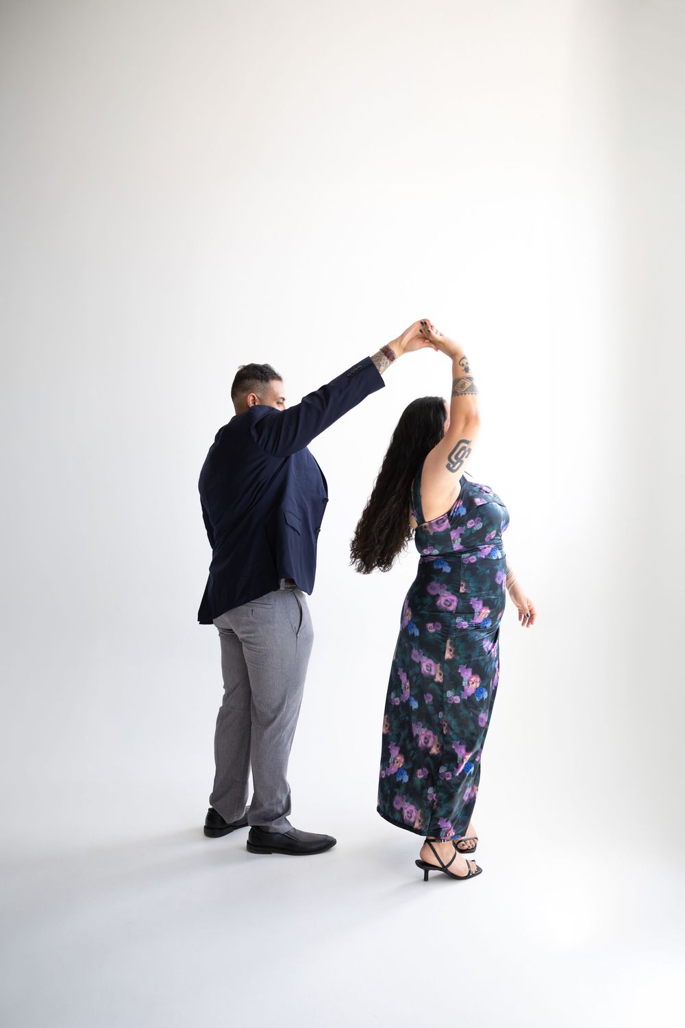 Couple dancing together, man in navy blazer and gray pants, woman in floral maxi dress, white studio background.