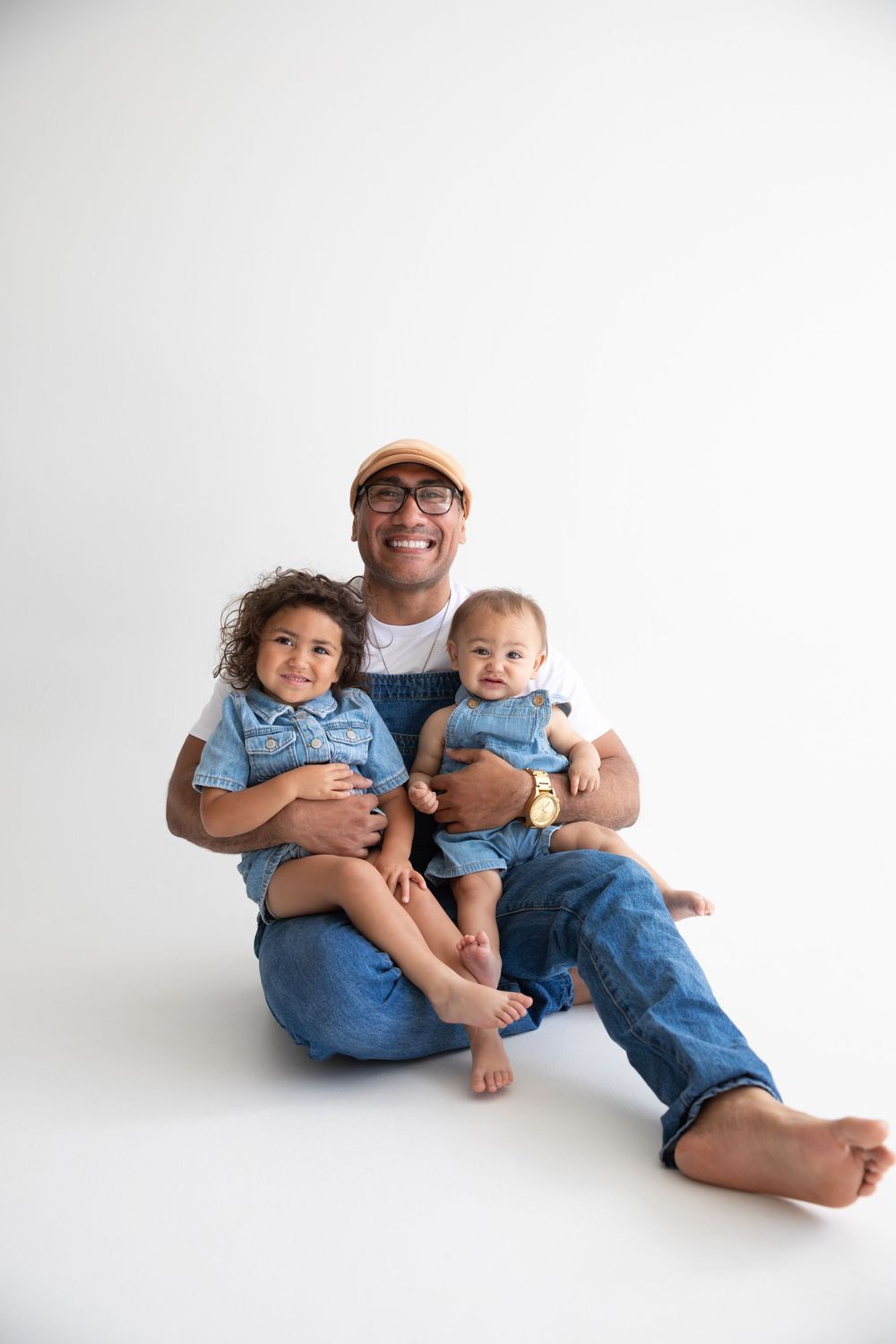 Smiling father sitting on white background holding two young children dressed in matching denim outfits.