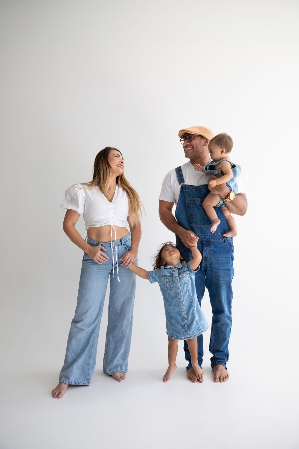 Family of four in denim outfits posing on white background, parents with two young children smiling.