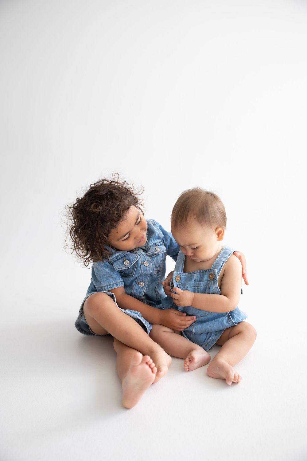 Two young siblings in matching denim outfits sit together on white background, older child lovingly watching baby.
