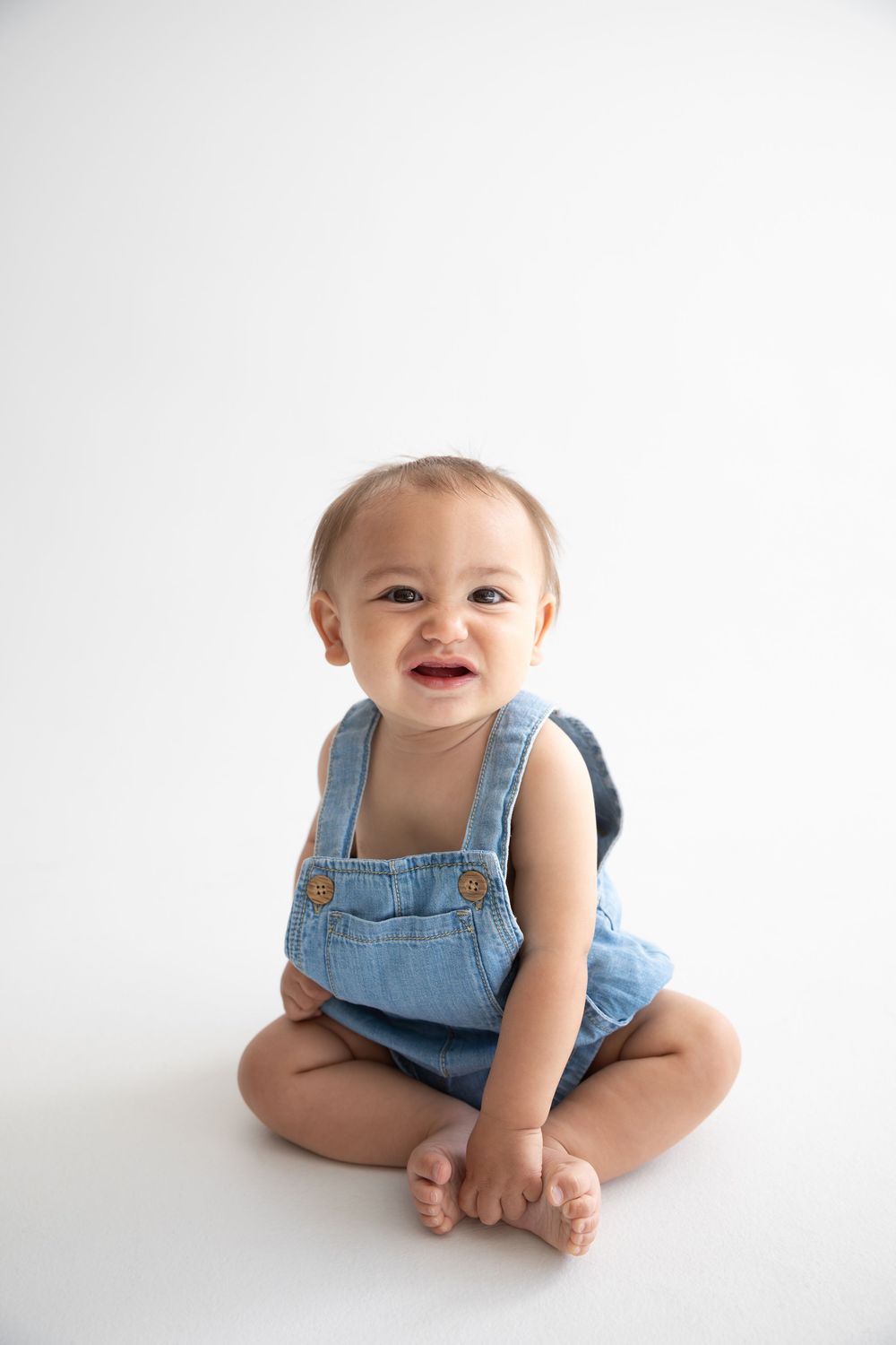 Smiling baby in denim overalls sitting cross-legged on white background.