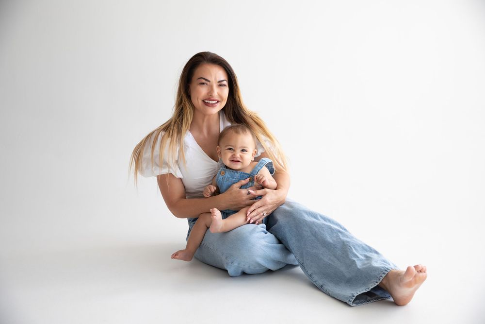 A smiling woman in white top and jeans sits on white floor holding a happy baby in blue outfit.
