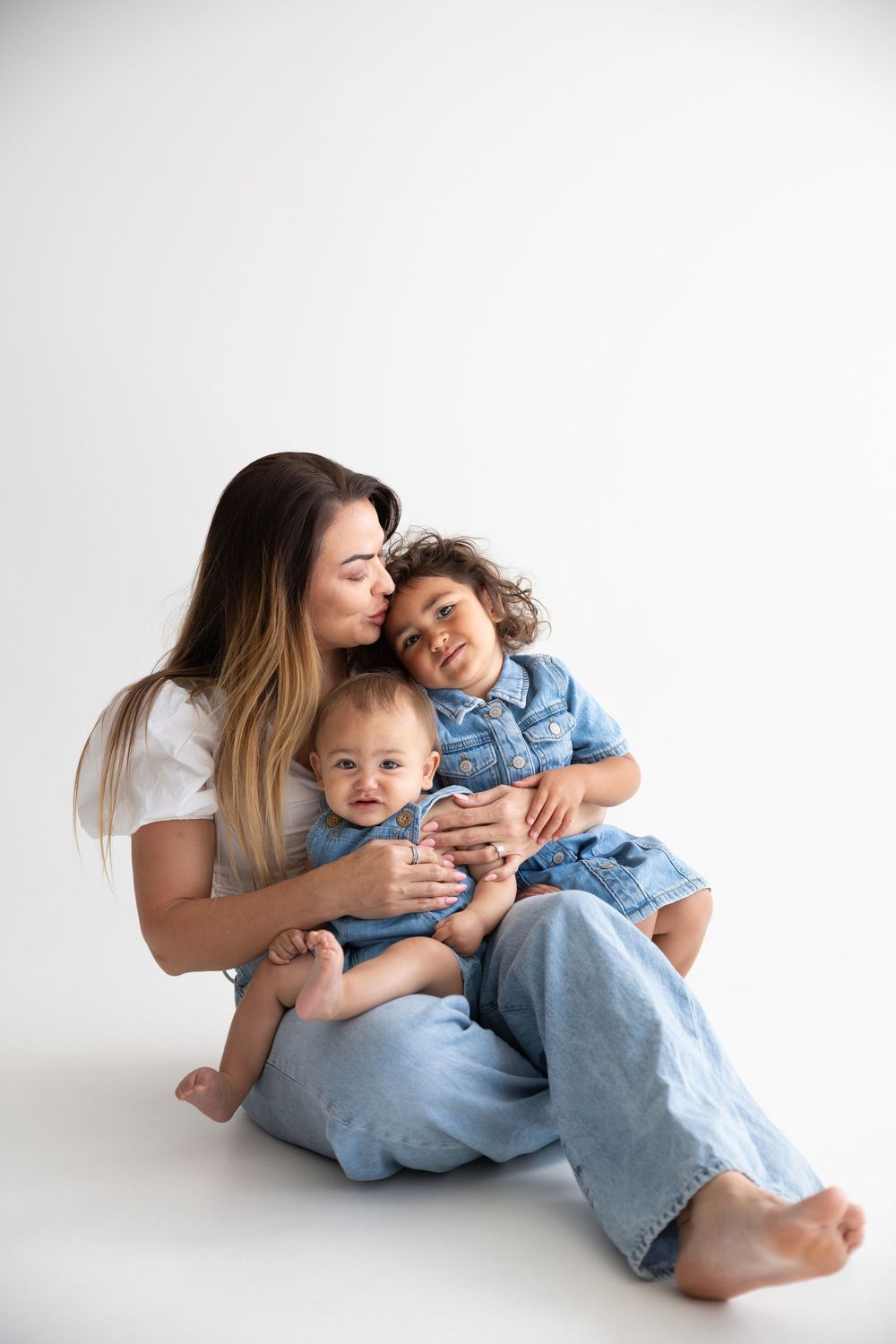 A mother sits on white background kissing her toddler while holding her smiling baby, all wearing denim outfits.