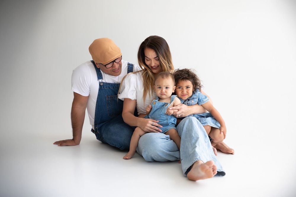 A happy family of four sitting together on a white background, parents with two young toddlers in denim outfits.