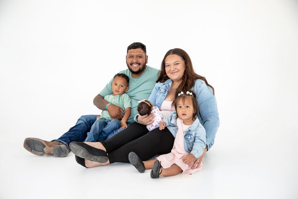 A happy family of four poses together on a white background, with two young children and smiling parents in casual attire.