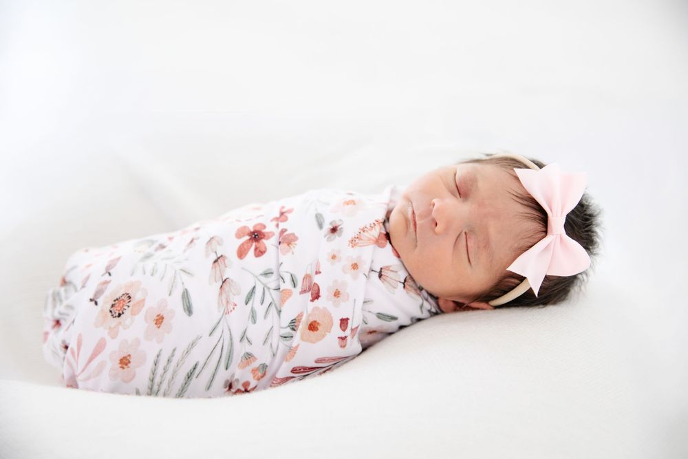 Sleeping newborn baby swaddled in a floral blanket with a pink bow headband on a white background.