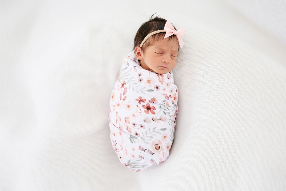 Newborn baby girl swaddled in floral wrap with pink bow headband, sleeping peacefully on white background.