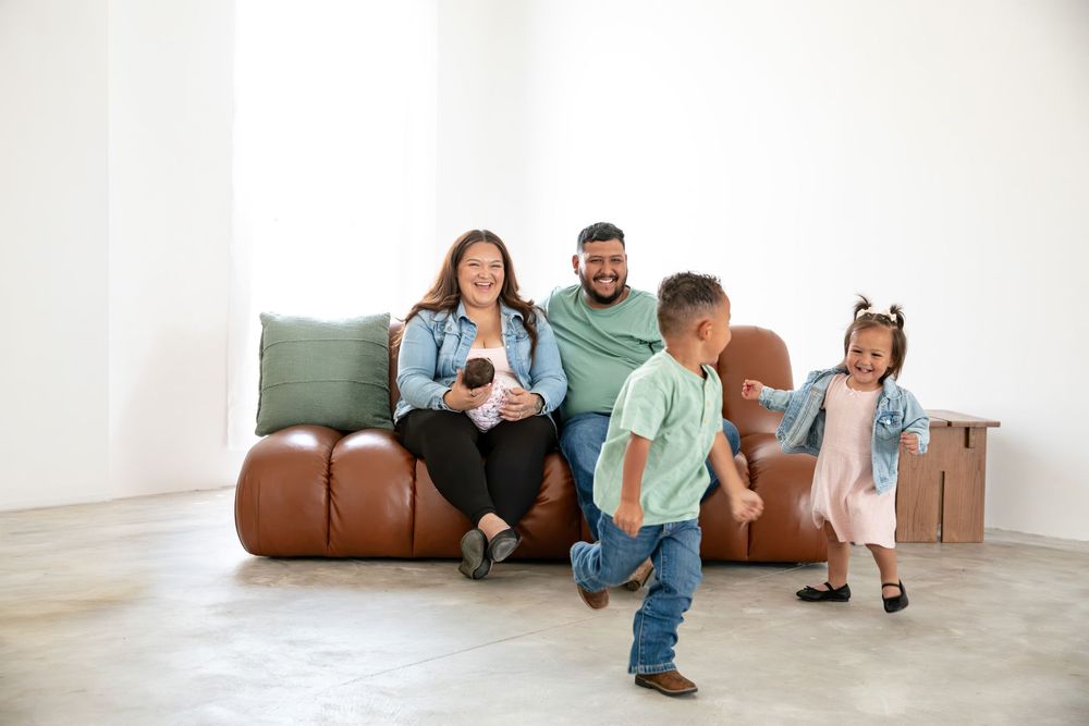 Happy family with parents on brown leather sofa watching two young children play in bright white living room.