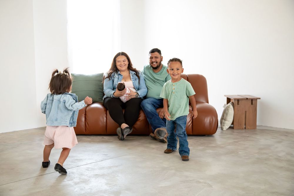 Happy family of four with newborn sitting on brown leather sofa in bright living room during home move.