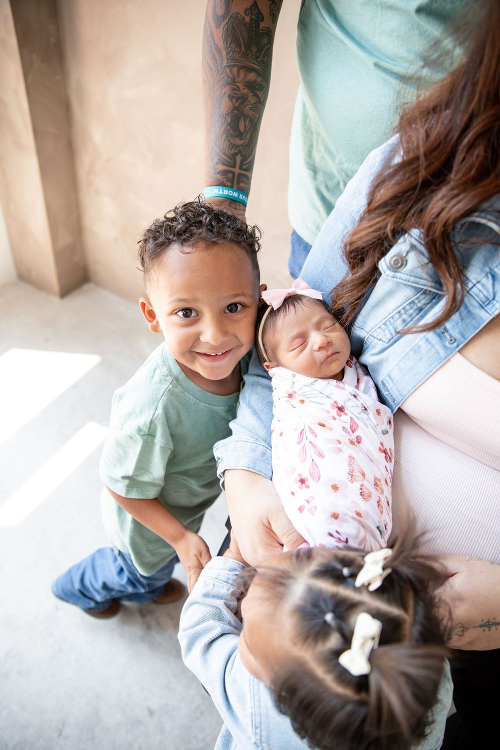 A mother holds a swaddled newborn while two young children gather close for a family portrait indoors.