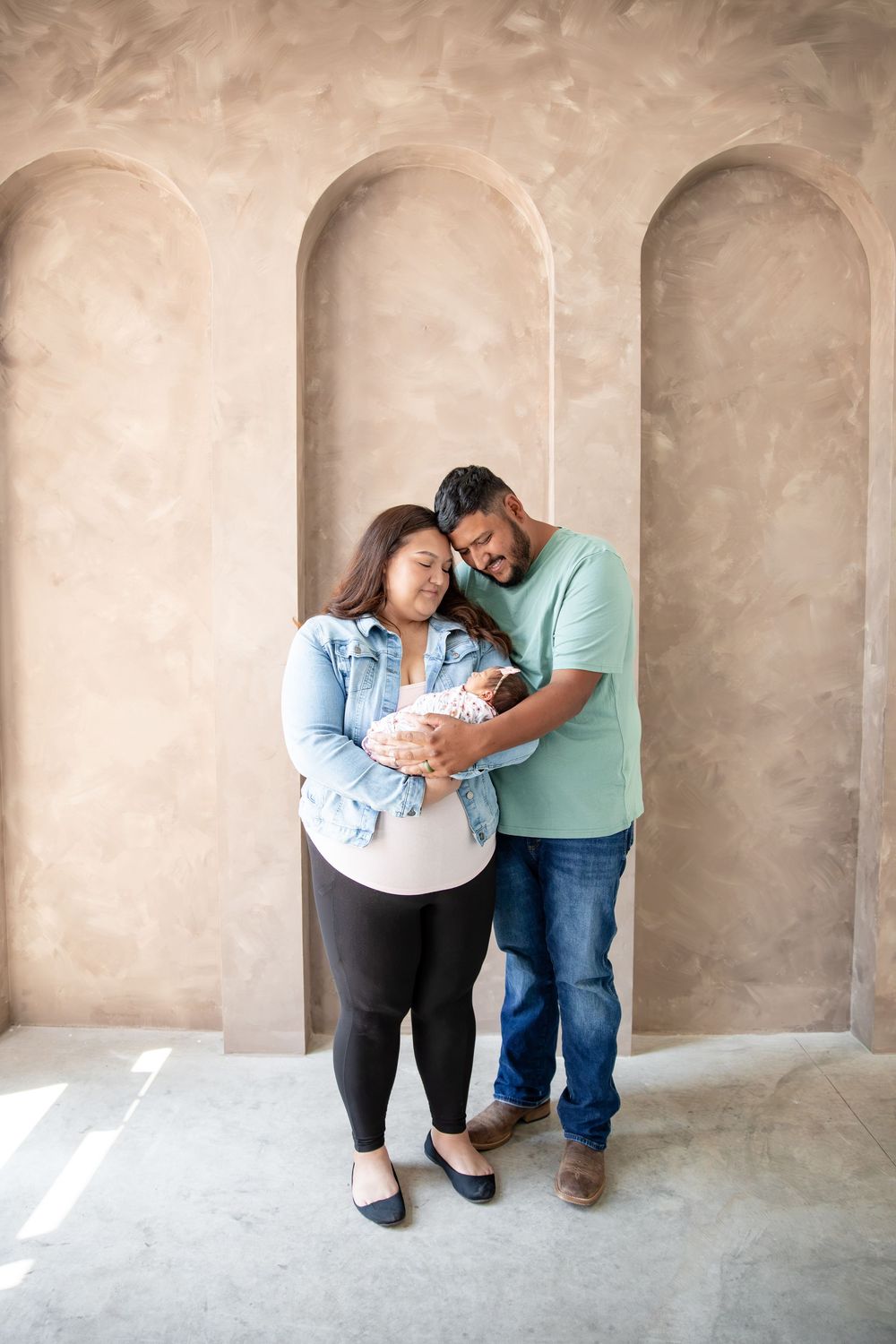 New parents tenderly cradle their newborn baby together, standing before elegant arched wall niches in a warm studio.