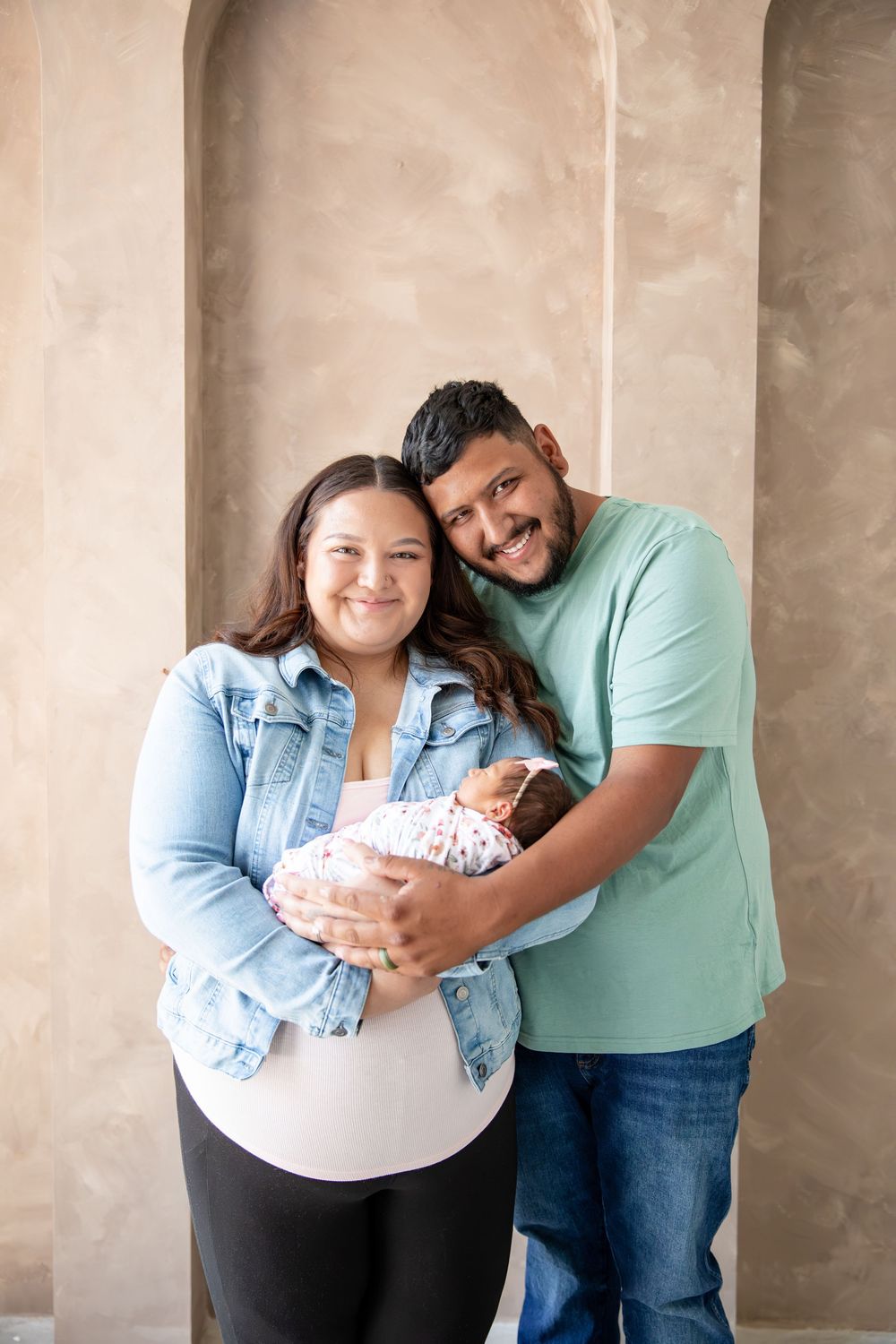 Happy couple smiling and holding their newborn baby, dressed casually, standing against a neutral beige wall.