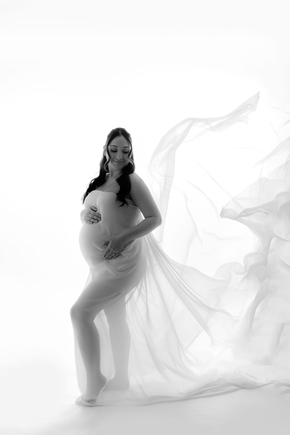 A flowing white maternity gown floats dramatically in a minimalist black and white studio photoshoot.